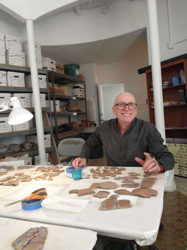 A man is sitting at a table in an archaeological laboratory. He is using blue masking tape to join pottery sherds that fit together.