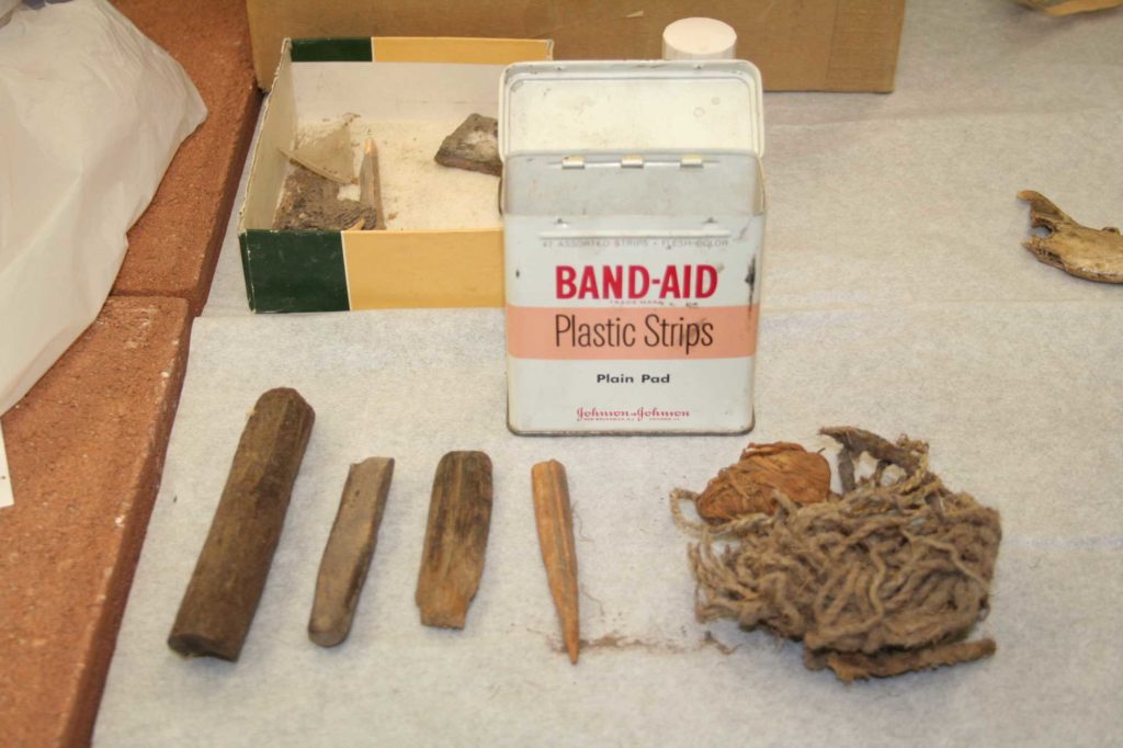 This image shows ancient perishable objects that have been removed from the collector's storage container. In the foreground at left are four items made of animal bone. At right is a bundle of cordage made from plant fibers. In mid-ground is a 1950s or 1960s-era metal Johnson & Johnson Band-Aid box with its lid open. In the background is an open cardboard box lid with indeterminate archaeological objects.